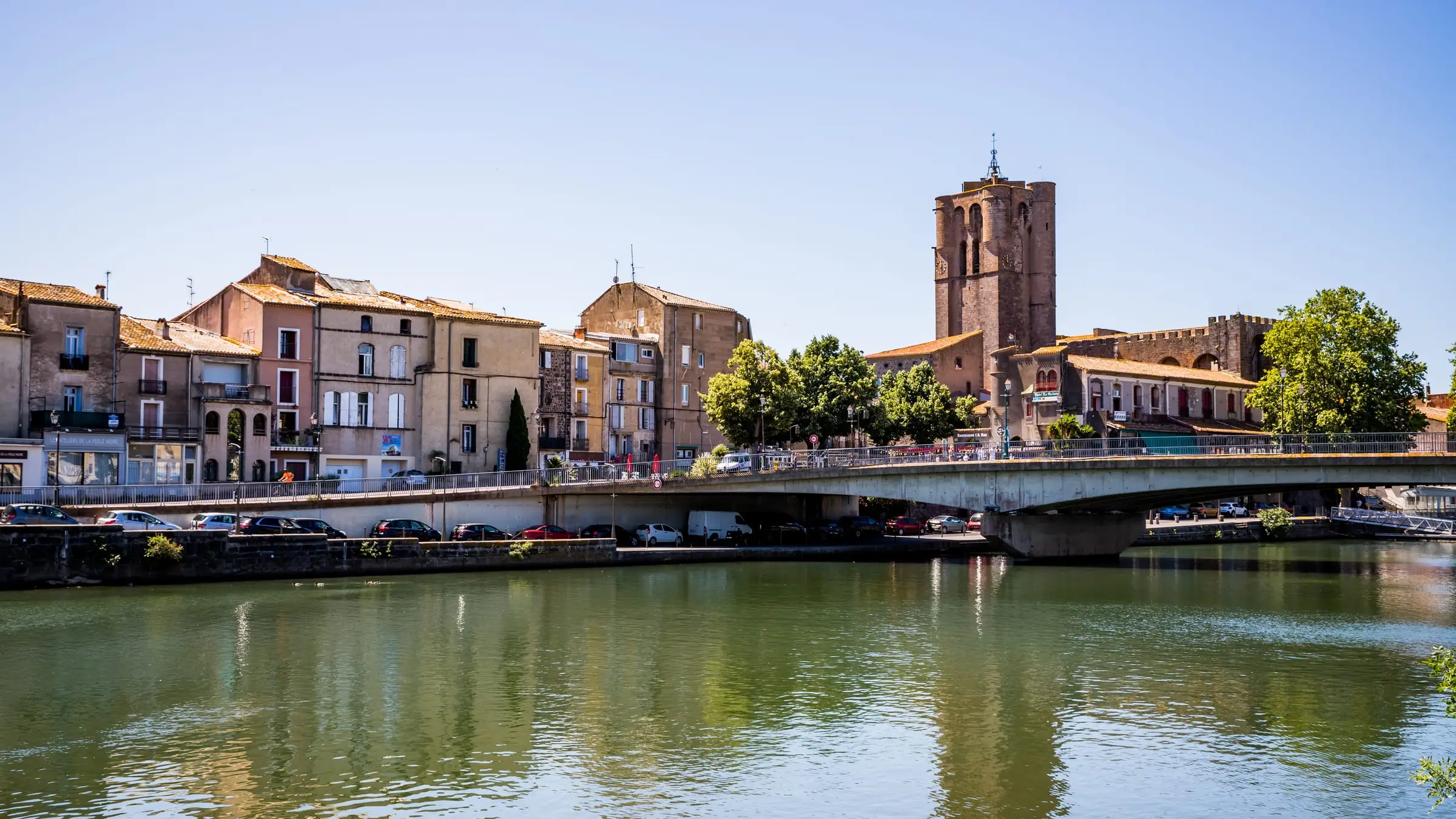 La cathédrale Saint-Étienne et les maisons des quais d’Agde se reflètent dans l’Hérault, sous un ciel d’été