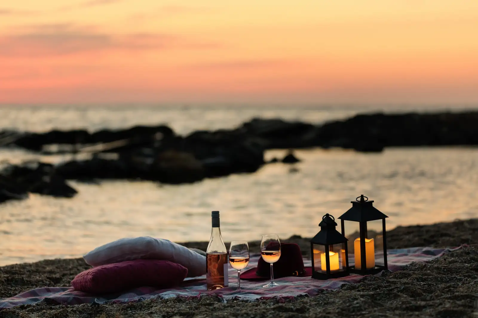 Deux verres de rosé, des coussins et des lanternes posés sur une plage au coucher du soleil