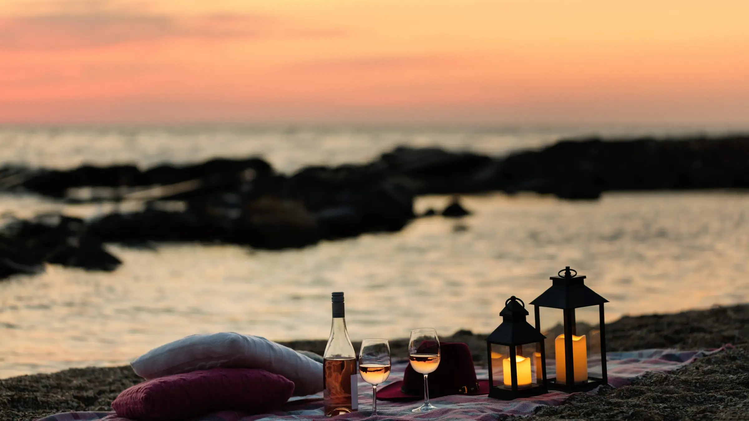 Deux verres de rosé, des coussins et des lanternes posés sur une plage au coucher du soleil