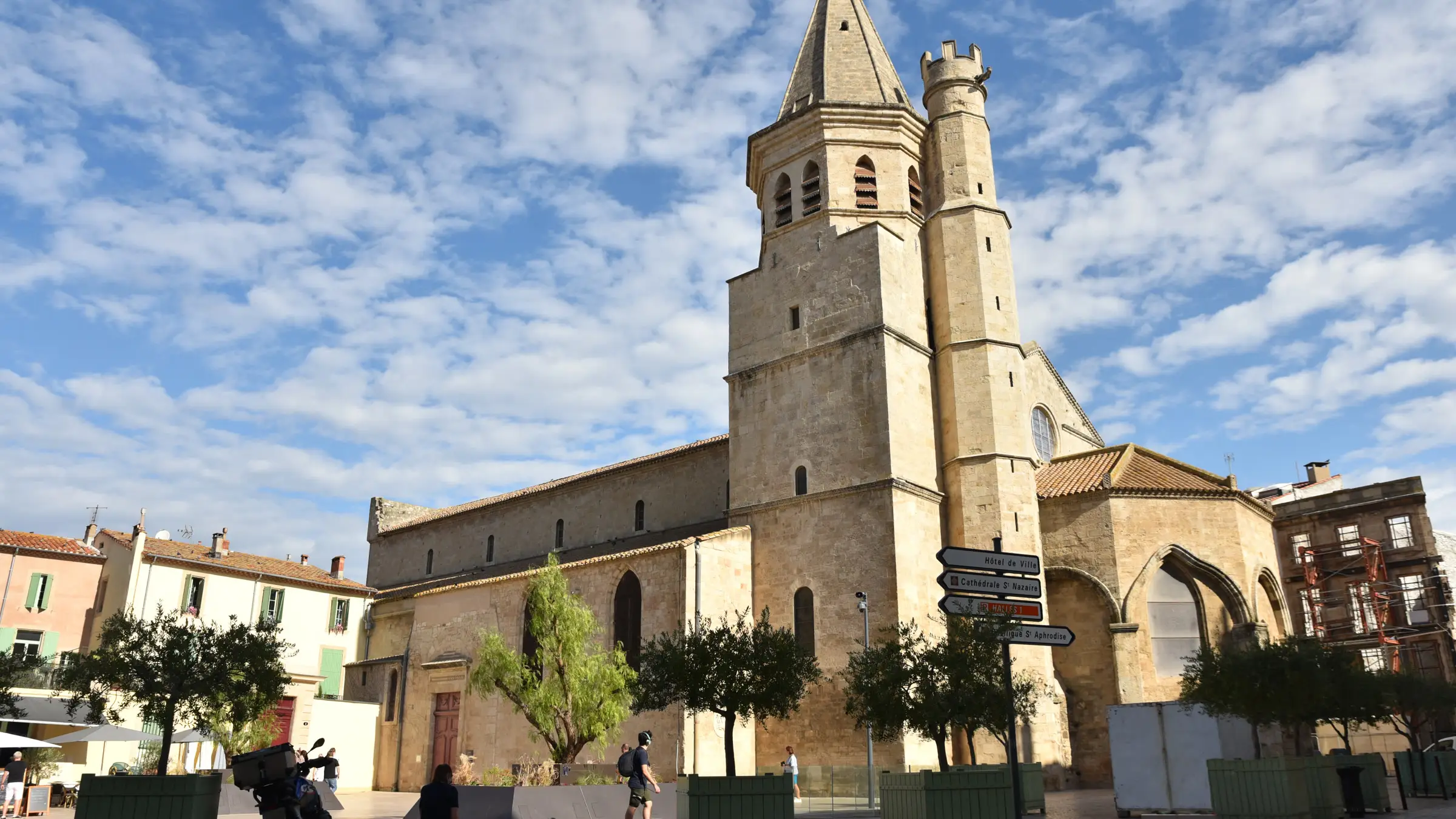 L’église de la Madeleine de Béziers, son clocher à flèche et sa tourelle crénelée, sous un ciel de nuages blancs, place de la Madeleine