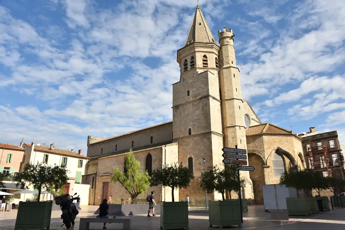 L’église de la Madeleine de Béziers, son clocher à flèche et sa tourelle crénelée, sous un ciel de nuages blancs, place de la Madeleine