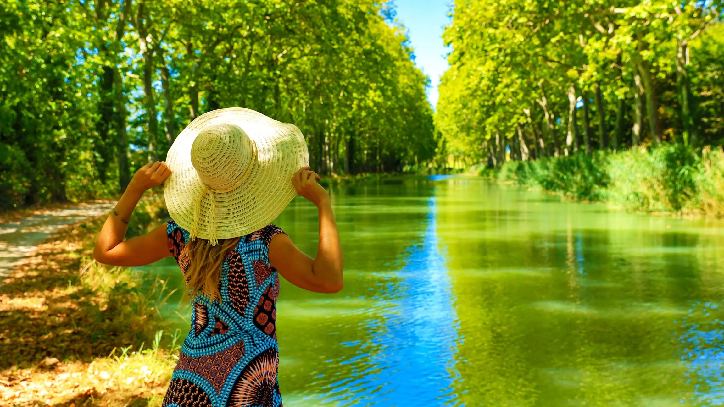 Une promeneuse en chapeau de paille, vue de dos, face au canal du Midi bordé de grands arbres