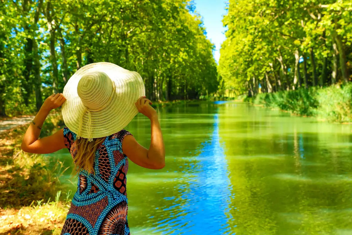 Une promeneuse en chapeau de paille, vue de dos, face au canal du Midi bordé de grands arbres