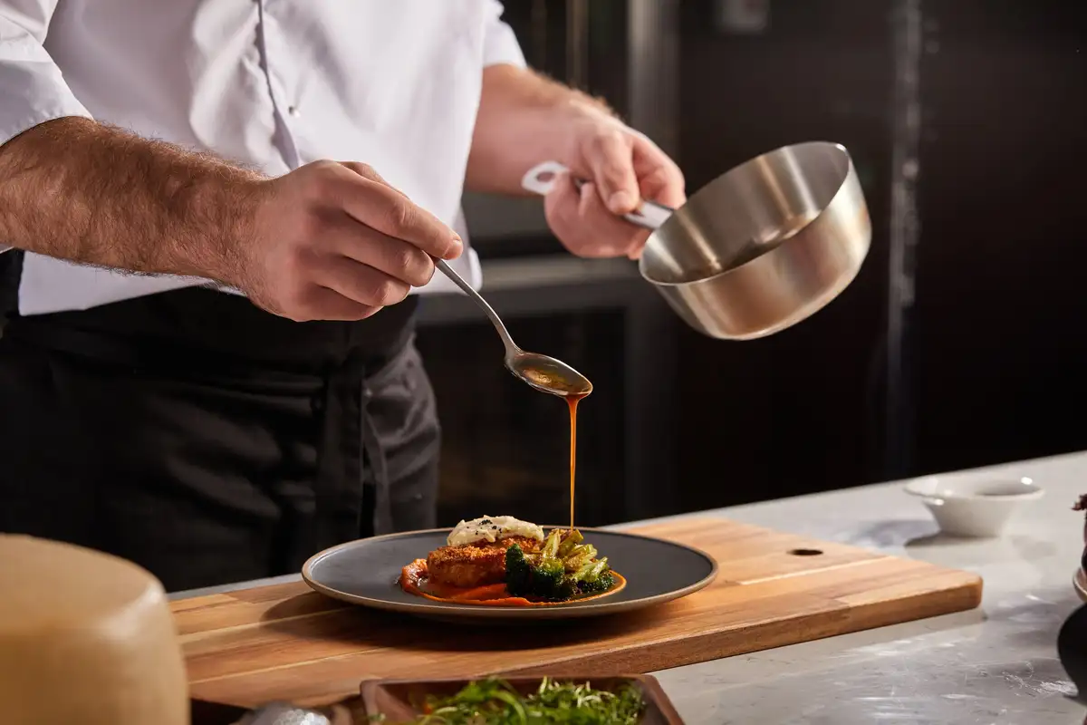 Un chef nappe de sauce une assiette dressée posée sur une planche en bois