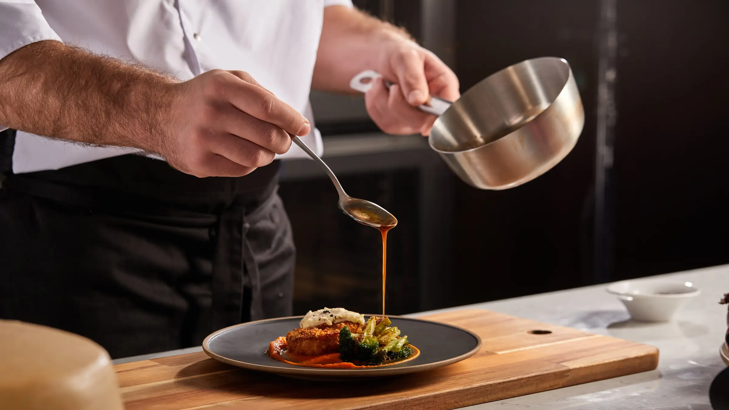 Un chef nappe de sauce une assiette dressée posée sur une planche en bois