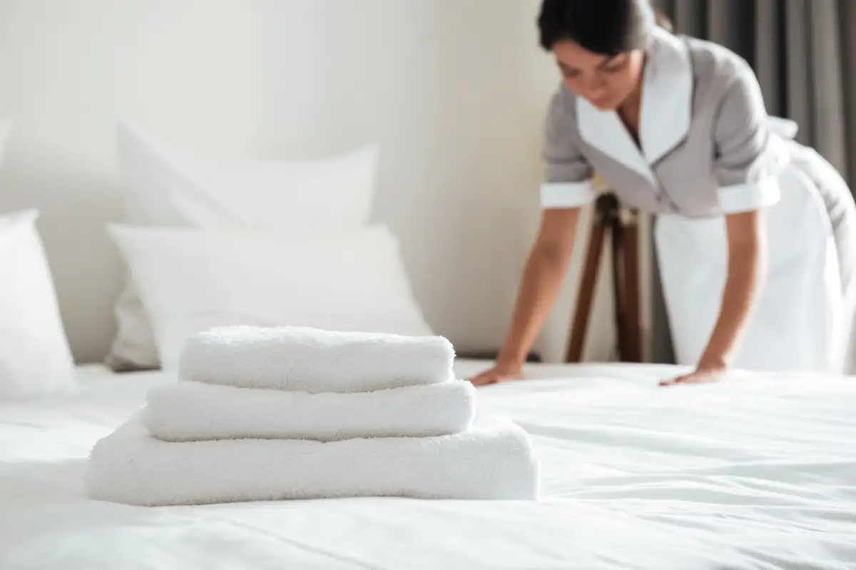 Serviettes blanches pliées sur un lit fait, une femme de chambre lisse les draps en arrière-plan