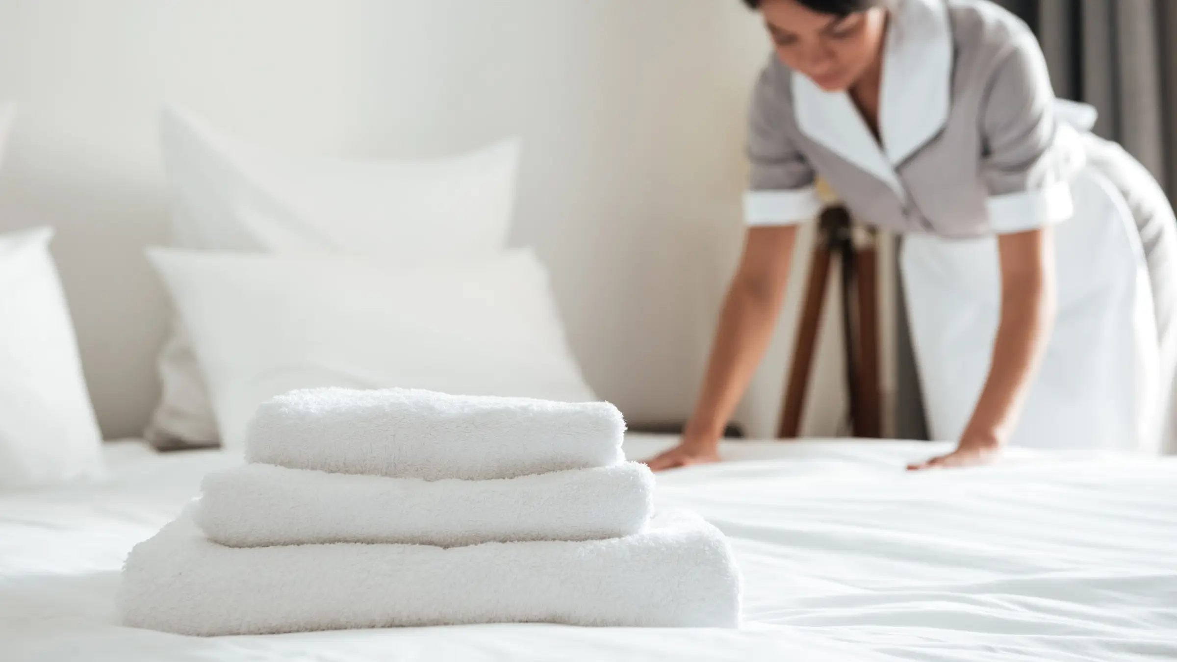 Serviettes blanches pliées sur un lit fait, une femme de chambre lisse les draps en arrière-plan