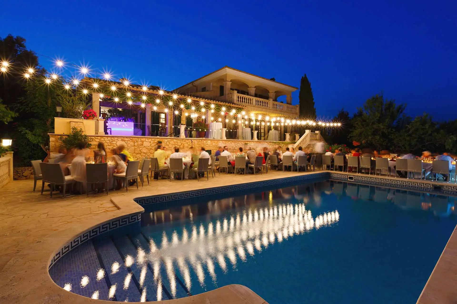 Dîner de fête au bord d’une piscine, à la nuit tombée, sous des guirlandes lumineuses