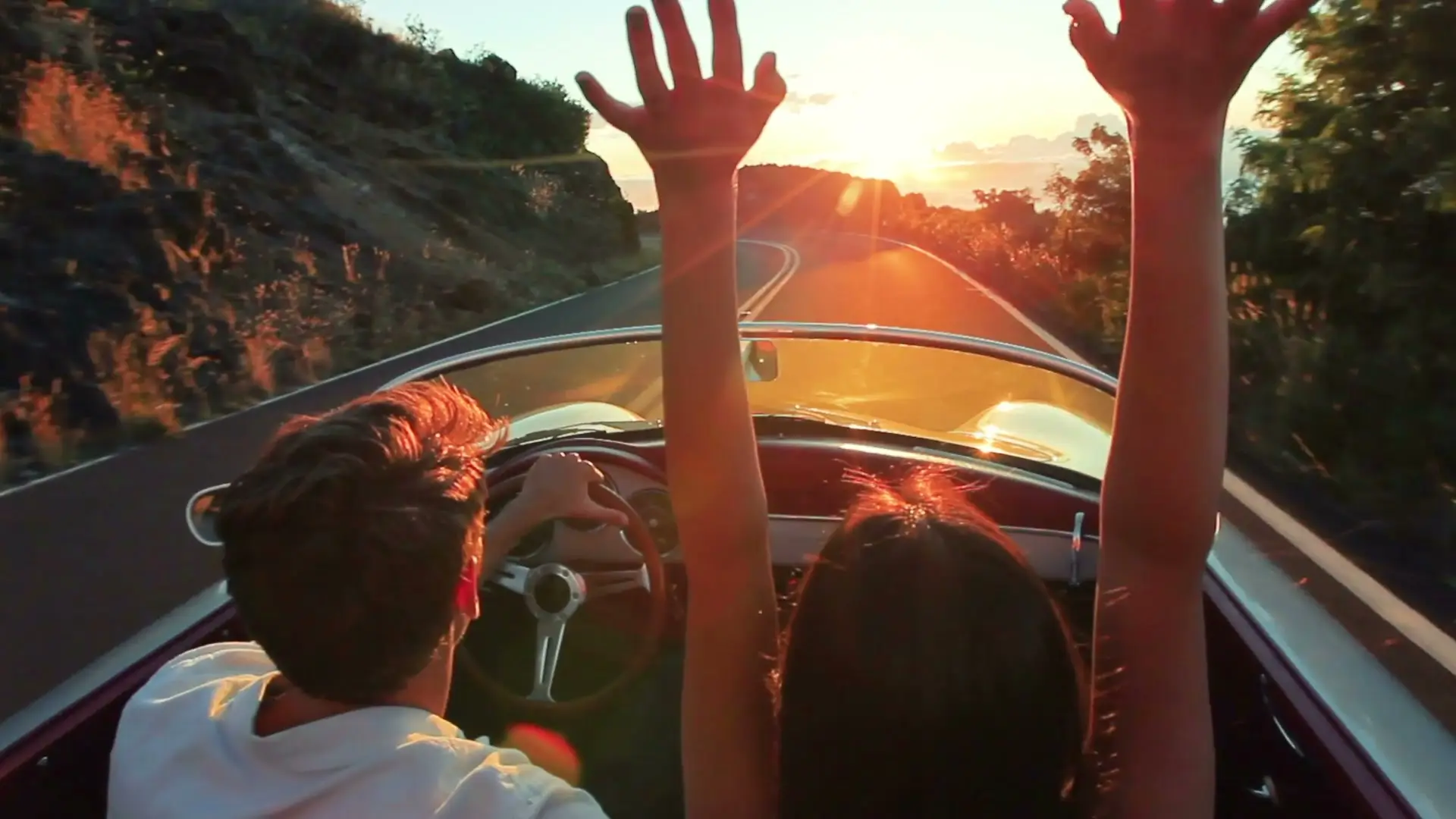Couple en cabriolet de collection, bras levés, sur une route côtière au coucher du soleil