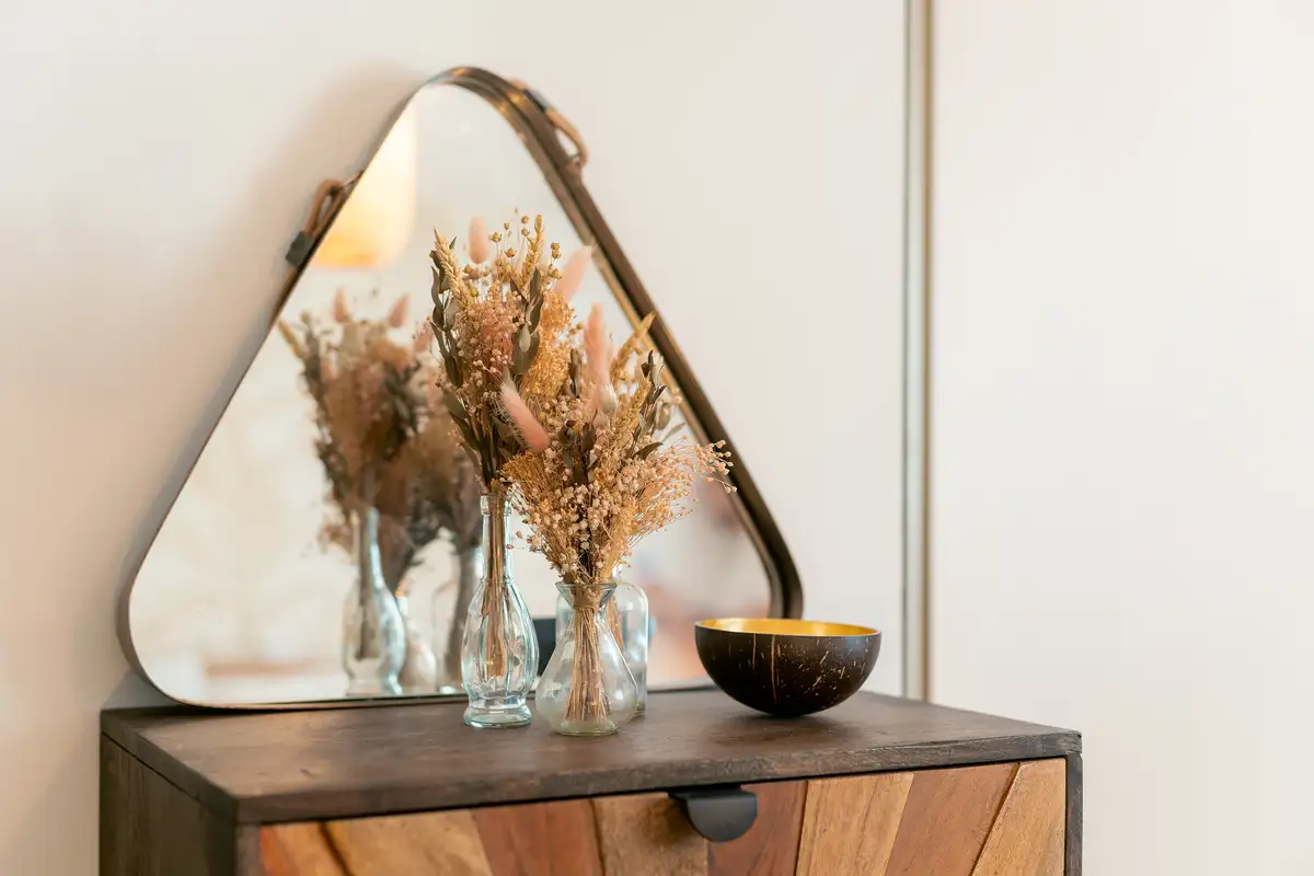 Un miroir aux angles arrondis, deux petits bouquets de fleurs séchées dans des vases de verre et une coupelle dorée sur une commode en bois