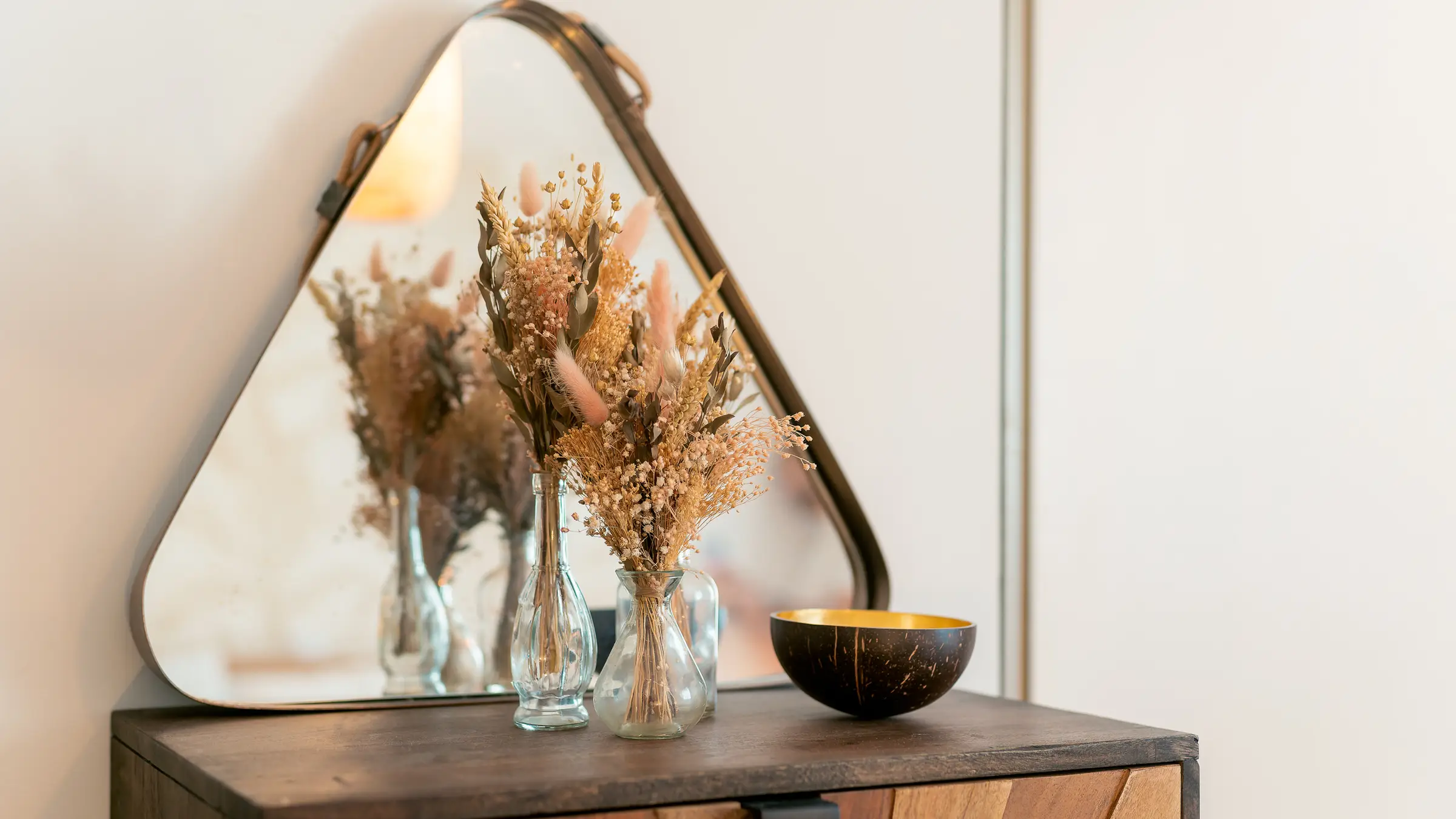 Un miroir aux angles arrondis, deux petits bouquets de fleurs séchées dans des vases de verre et une coupelle dorée sur une commode en bois
