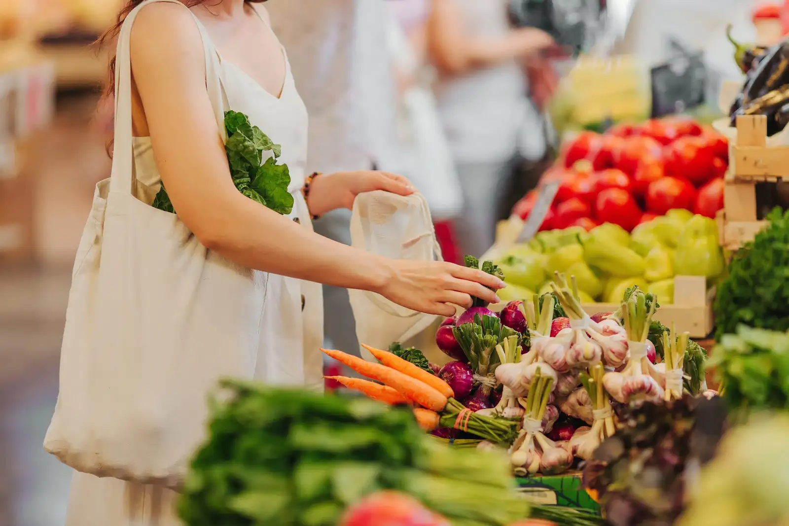 Au marché, une femme choisit des oignons rouges sur un étal de carottes, d’ail et de tomates, un panier en toile à l’épaule