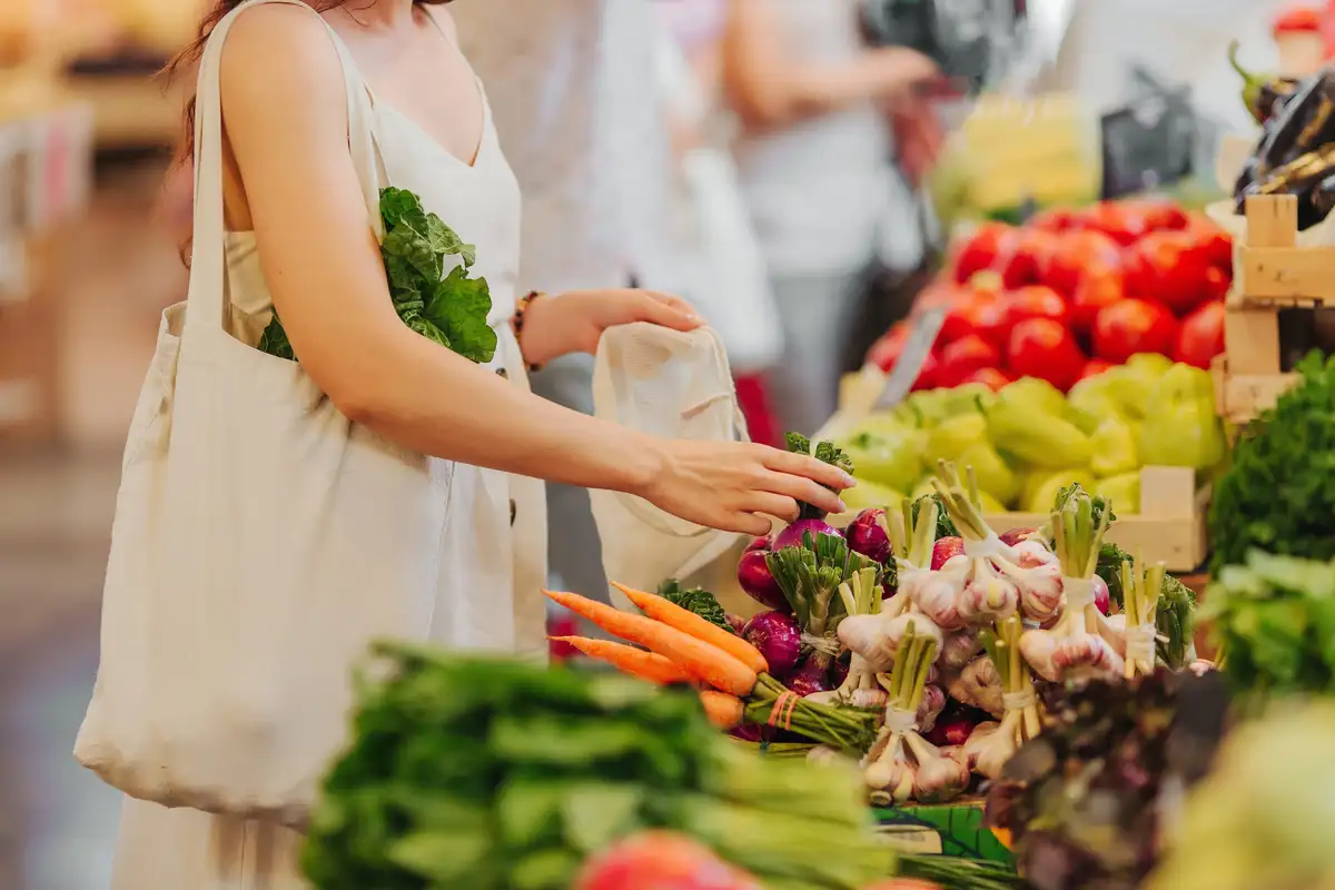 Au marché, une femme choisit des oignons rouges sur un étal de carottes, d’ail et de tomates, un panier en toile à l’épaule