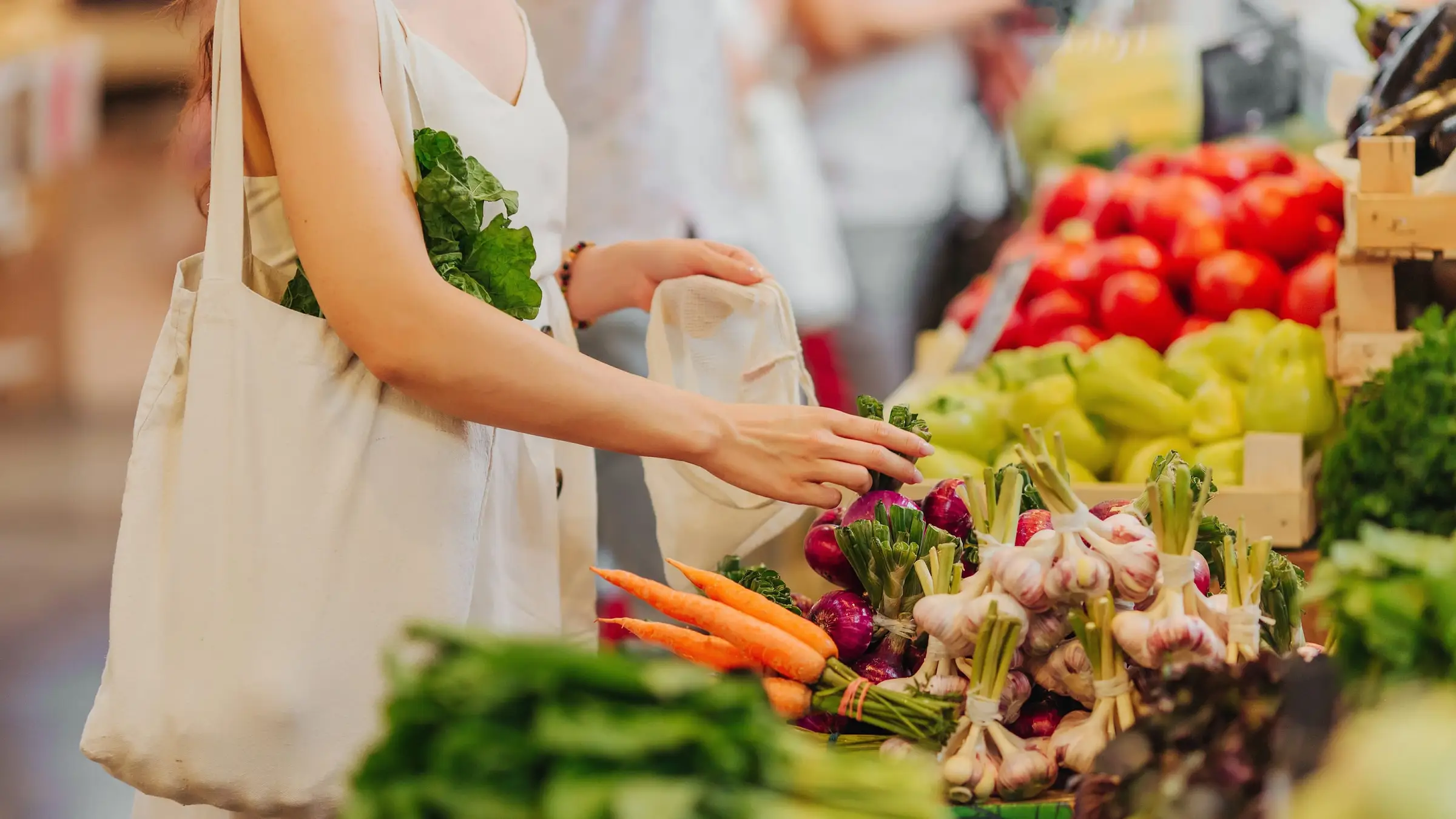 Au marché, une femme choisit des oignons rouges sur un étal de carottes, d’ail et de tomates, un panier en toile à l’épaule
