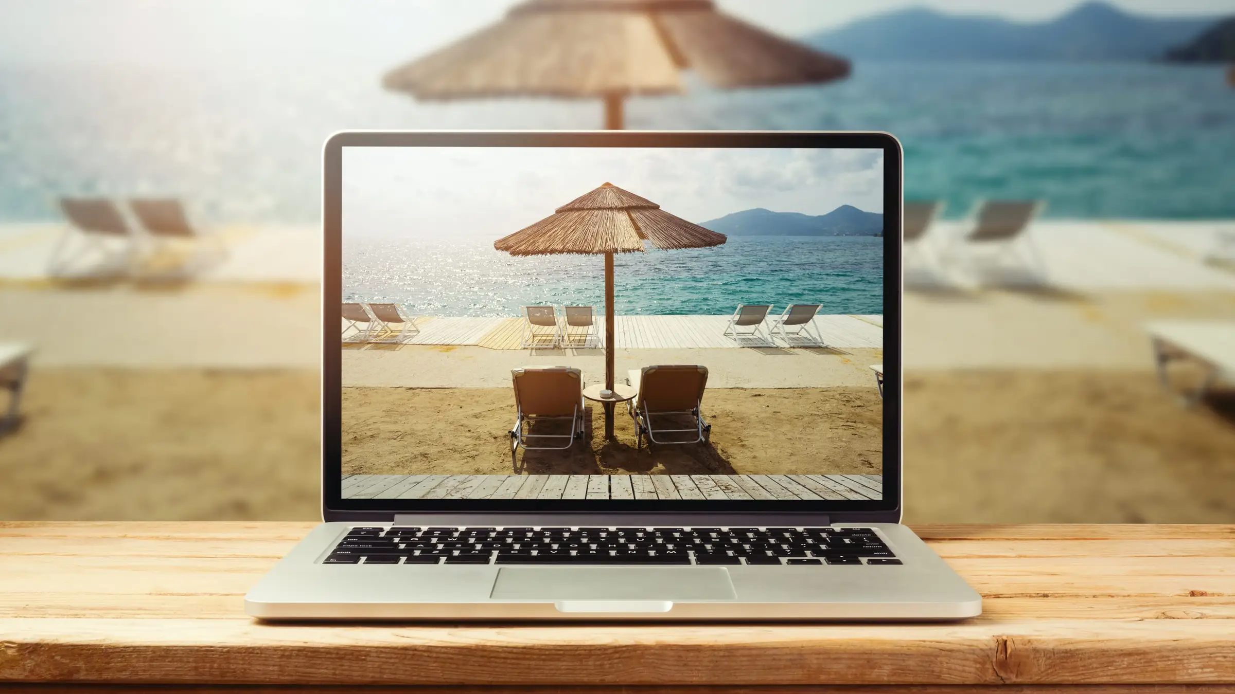 Un ordinateur portable posé sur une table en bois, dont l’écran montre une plage, un parasol de paille et deux transats