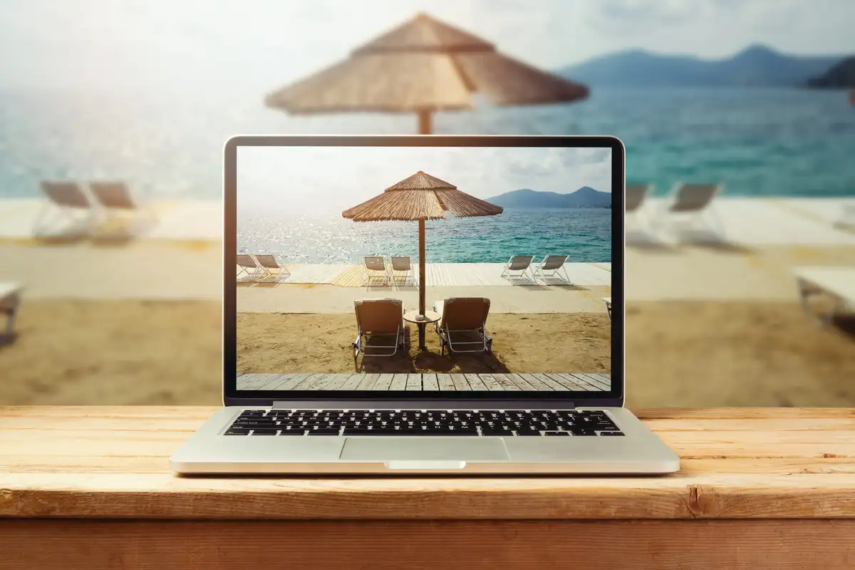 Un ordinateur portable posé sur une table en bois, dont l’écran montre une plage, un parasol de paille et deux transats