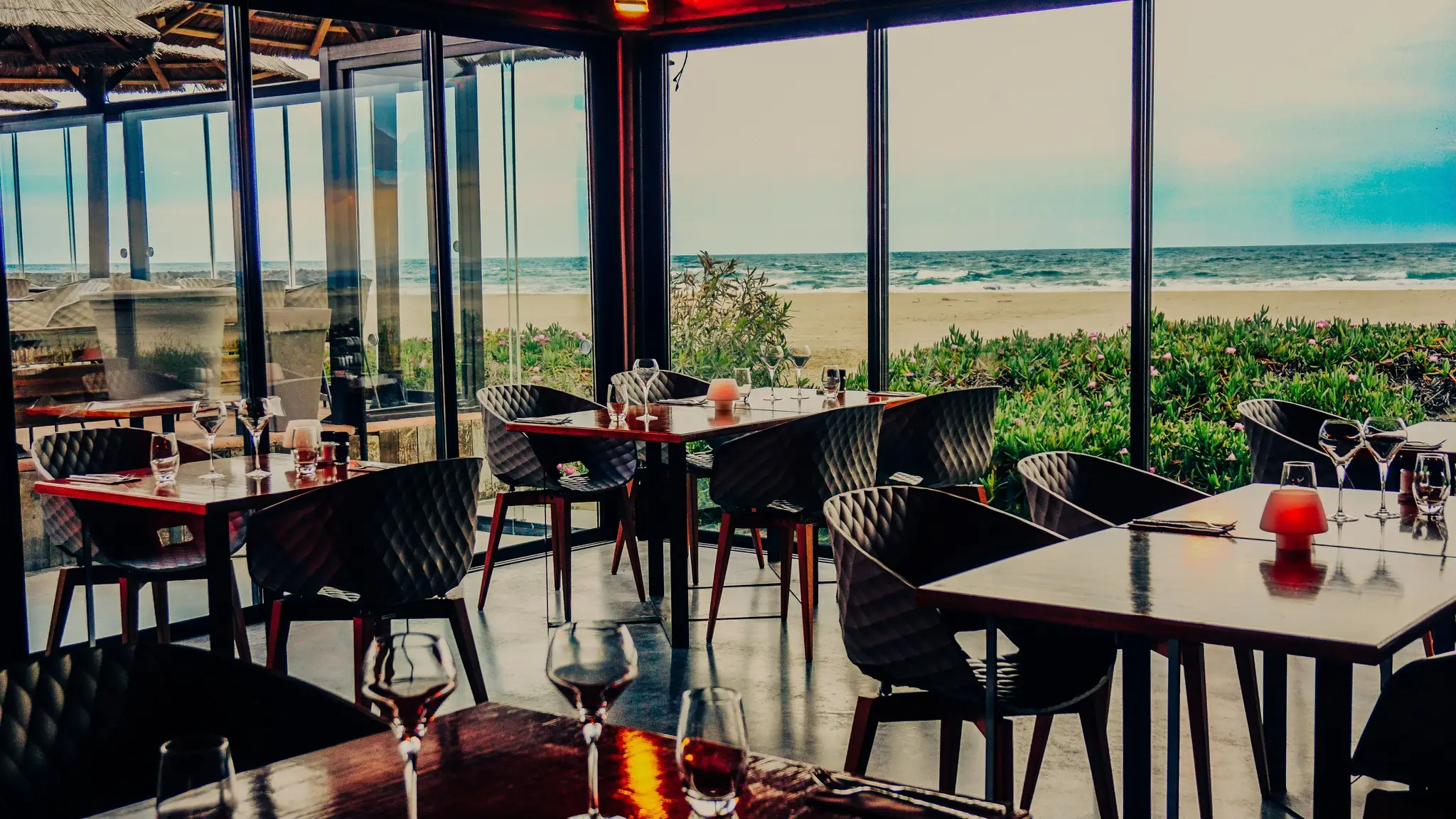 Salle du restaurant Les Vagues au Grau d’Agde, tables dressées derrière de grandes baies vitrées ouvertes sur la plage et la mer