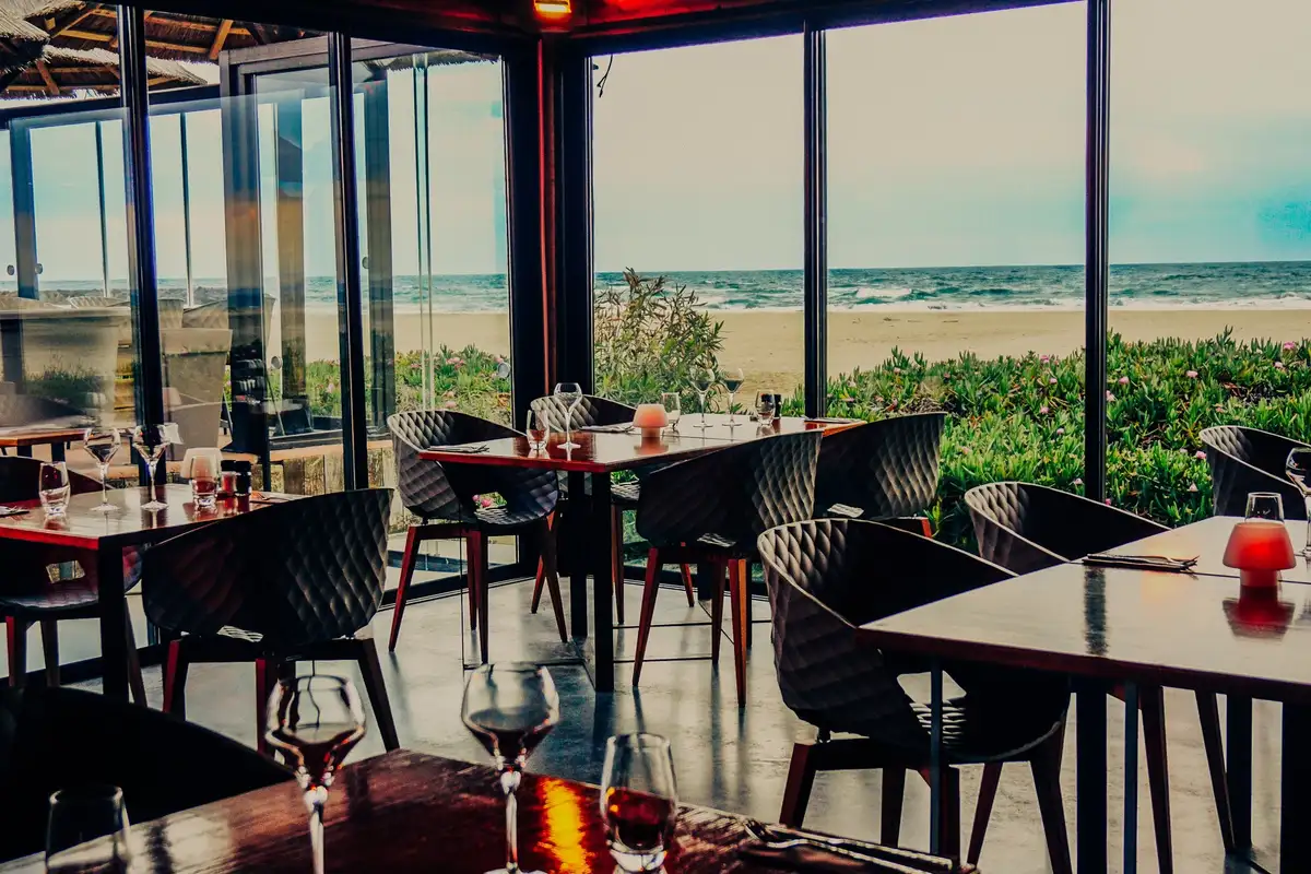 Salle du restaurant Les Vagues au Grau d’Agde, tables dressées derrière de grandes baies vitrées ouvertes sur la plage et la mer