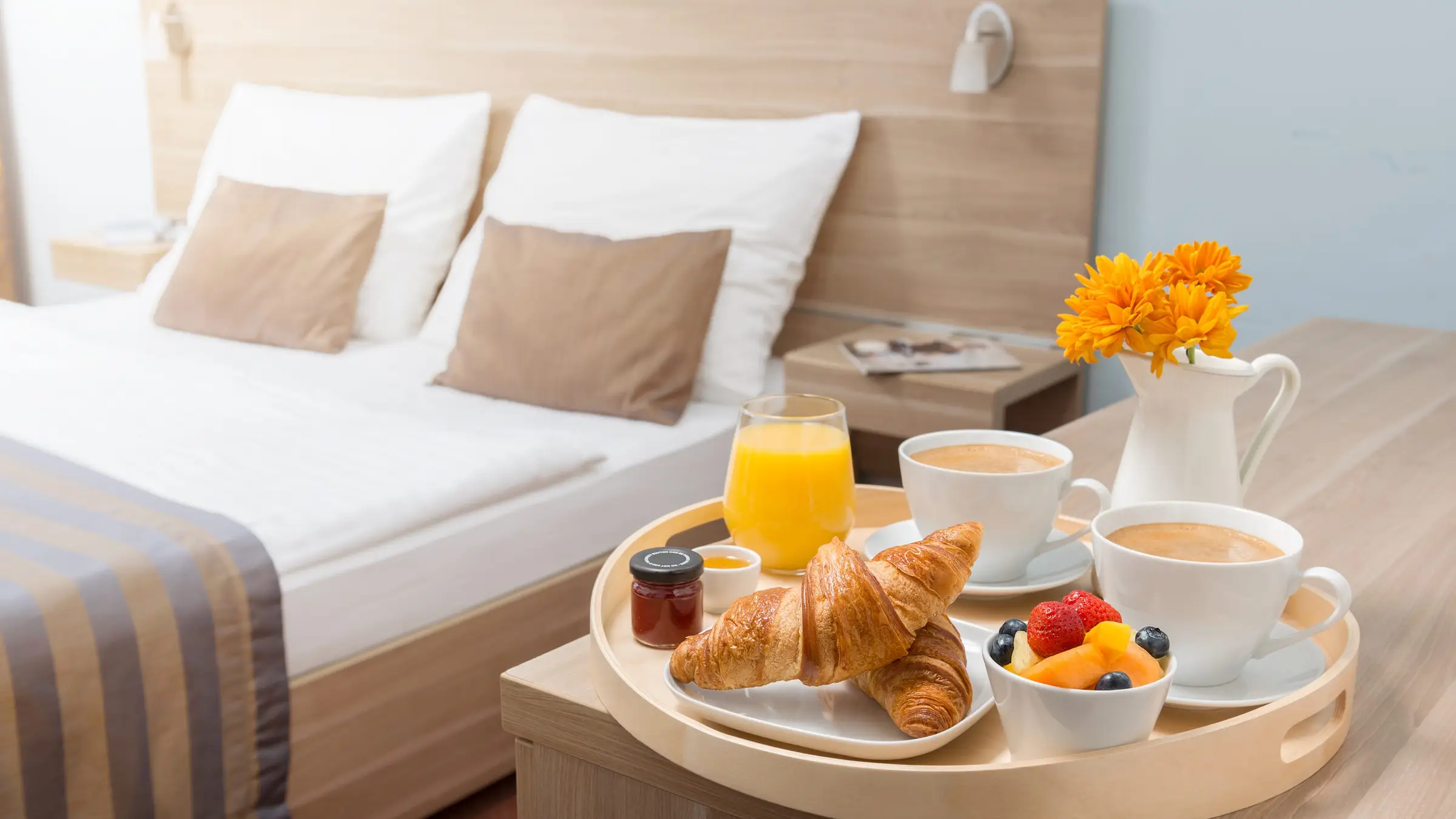 Plateau de petit-déjeuner posé près d’un lit : croissants, jus d’orange, café et corbeille de fruits