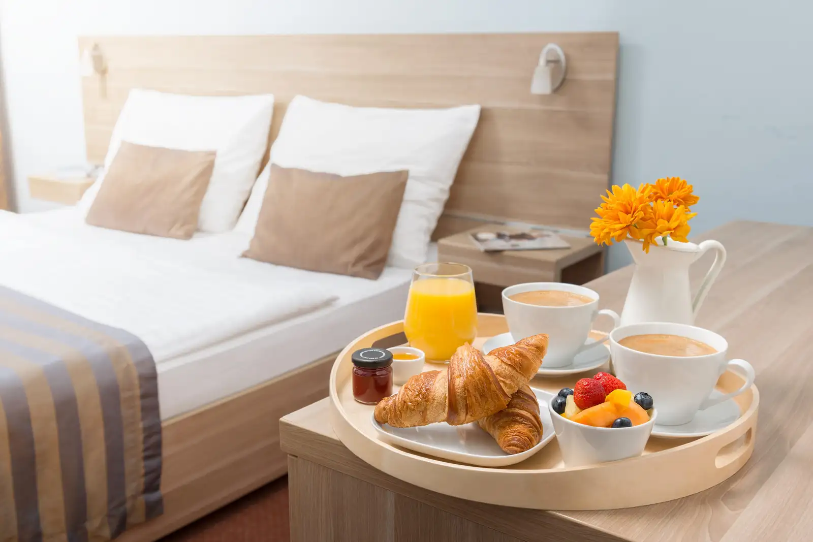 Plateau de petit-déjeuner posé près d’un lit : croissants, jus d’orange, café et corbeille de fruits