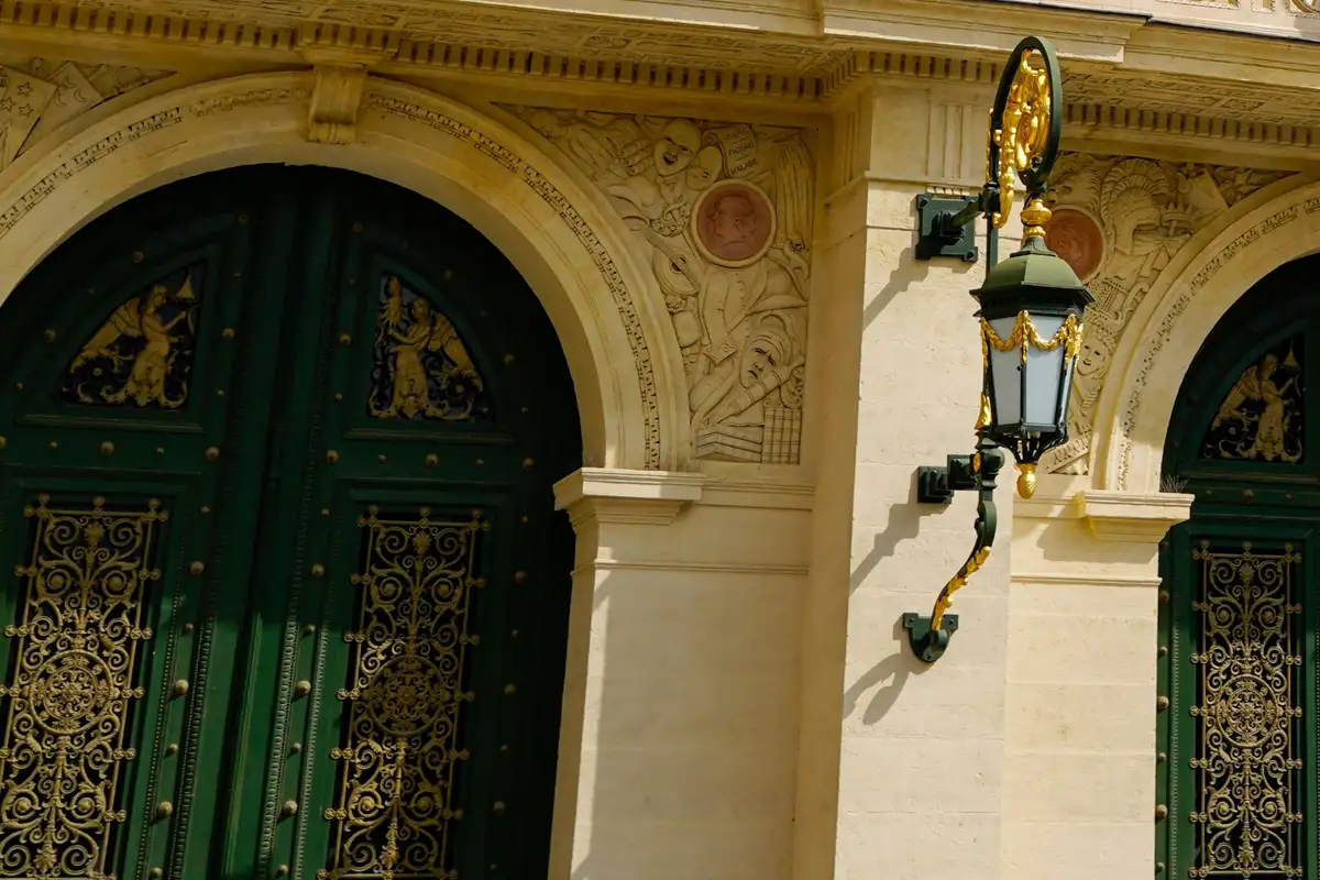 Hautes portes vertes à ferronneries dorées sous des arches de pierre sculptée, et une lanterne en fer forgé