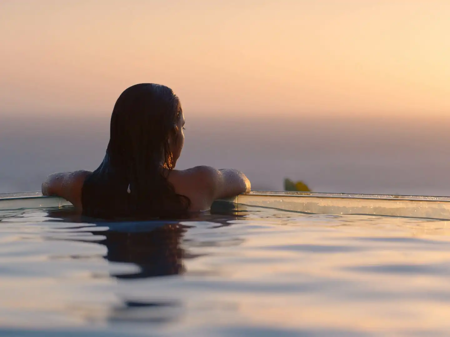 Une femme accoudée au bord d’une piscine à débordement contemple la mer au coucher du soleil