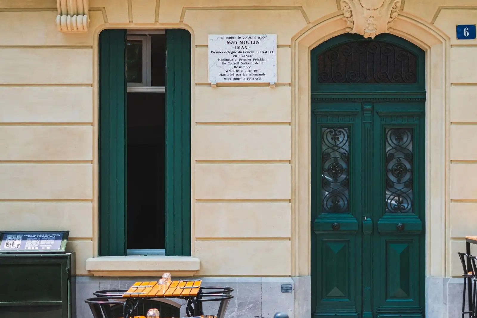 Façade de la Cave Chez Max à Béziers : porte et volets verts, plaque rappelant la naissance de Jean Moulin dans la maison, tables de la terrasse