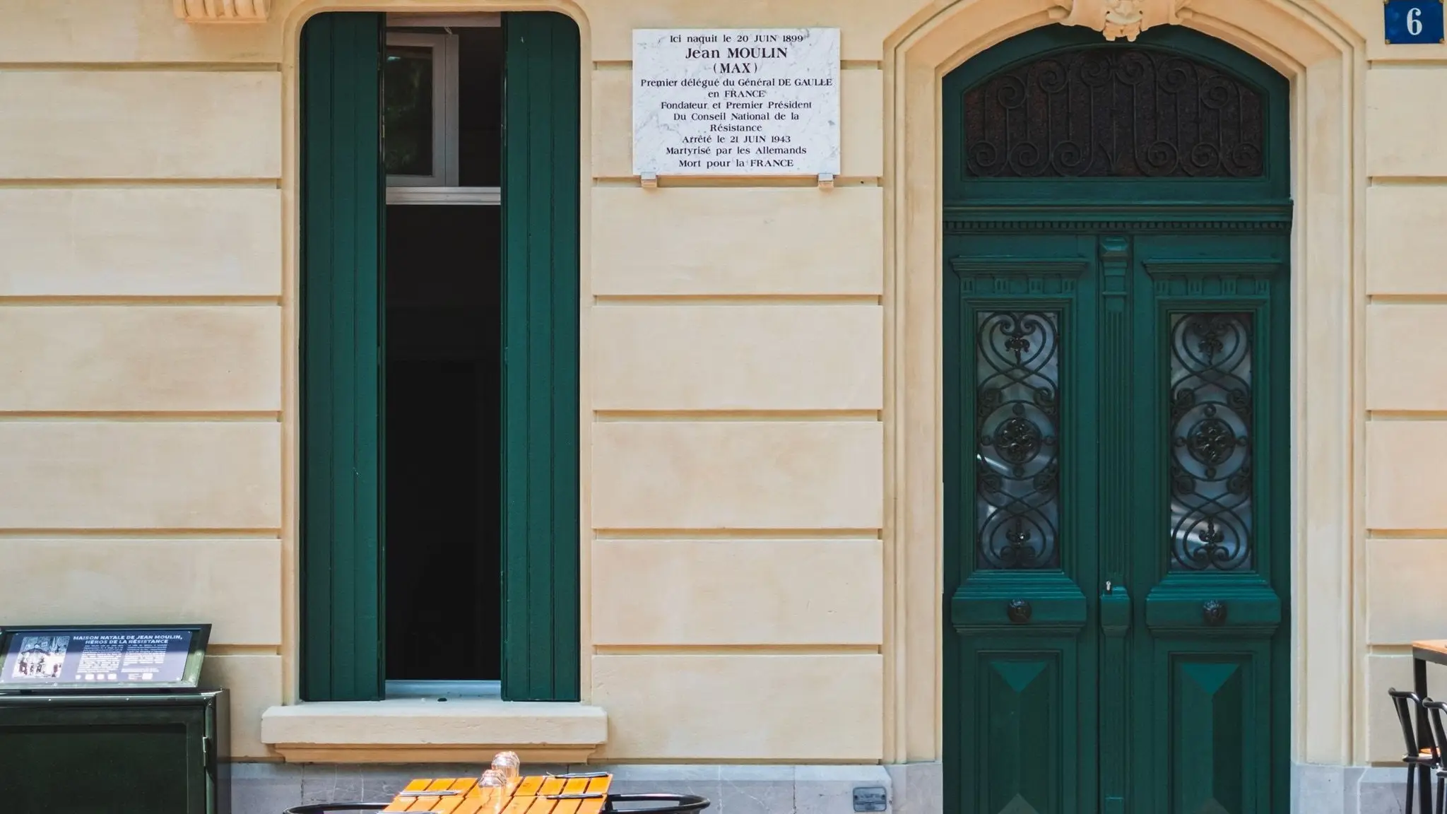 Façade de la Cave Chez Max à Béziers : porte et volets verts, plaque rappelant la naissance de Jean Moulin dans la maison, tables de la terrasse