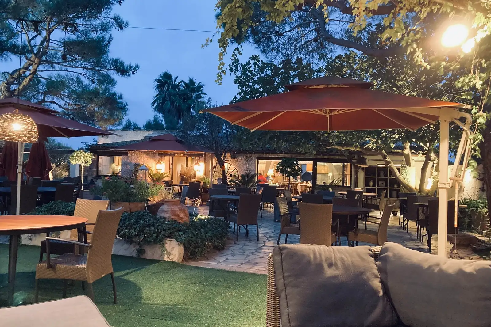 Terrasse arborée de Ma Ferme à Sérignan à la tombée de la nuit : tables sous les pins, parasols rouge brique et lumières suspendues, salle éclairée au fond