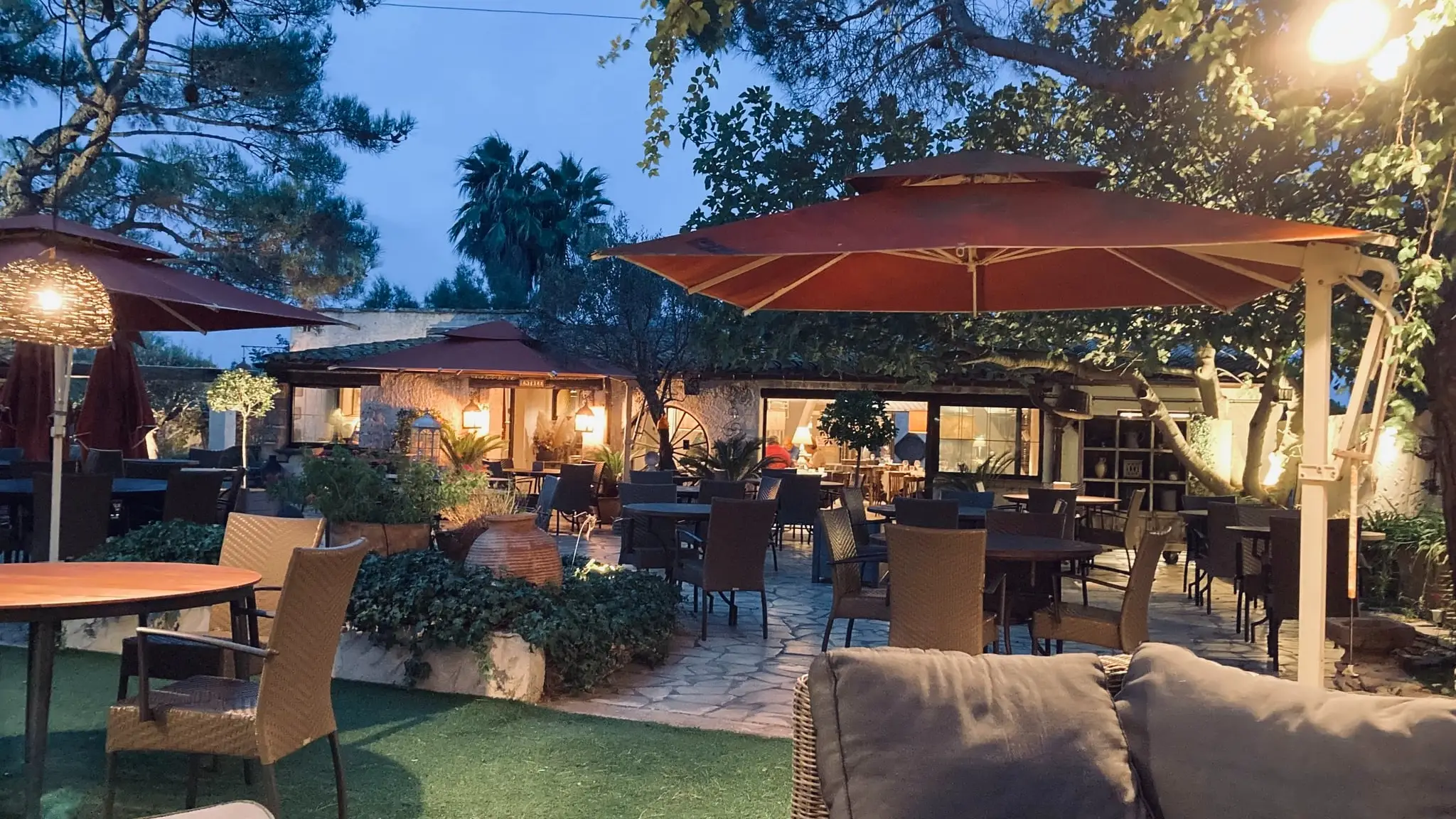 Terrasse arborée de Ma Ferme à Sérignan à la tombée de la nuit : tables sous les pins, parasols rouge brique et lumières suspendues, salle éclairée au fond