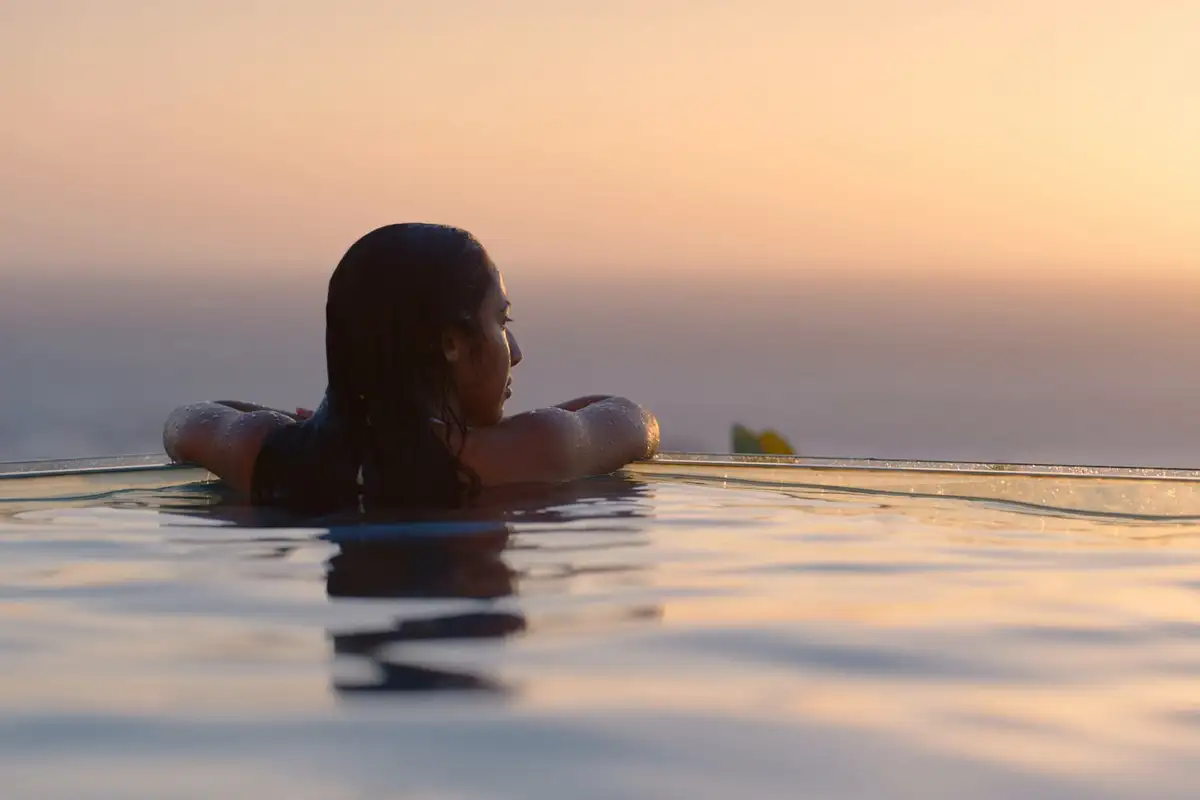De profil, une femme se repose au bord d’une piscine à débordement, face à la mer dans la lumière du soir
