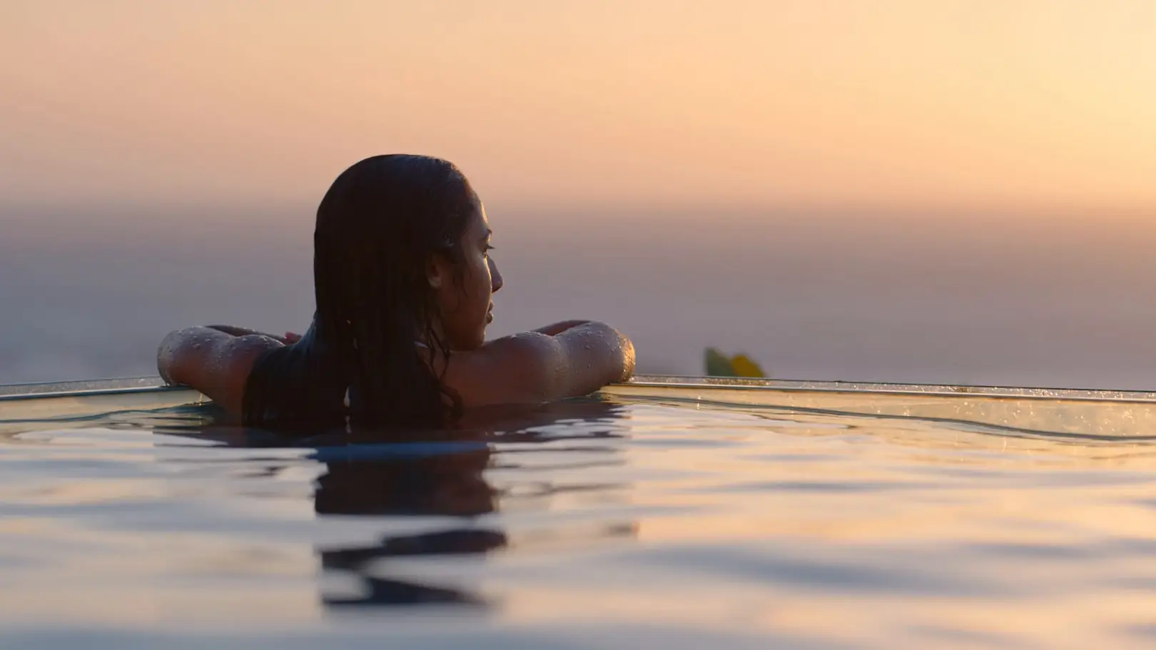 De profil, une femme se repose au bord d’une piscine à débordement, face à la mer dans la lumière du soir