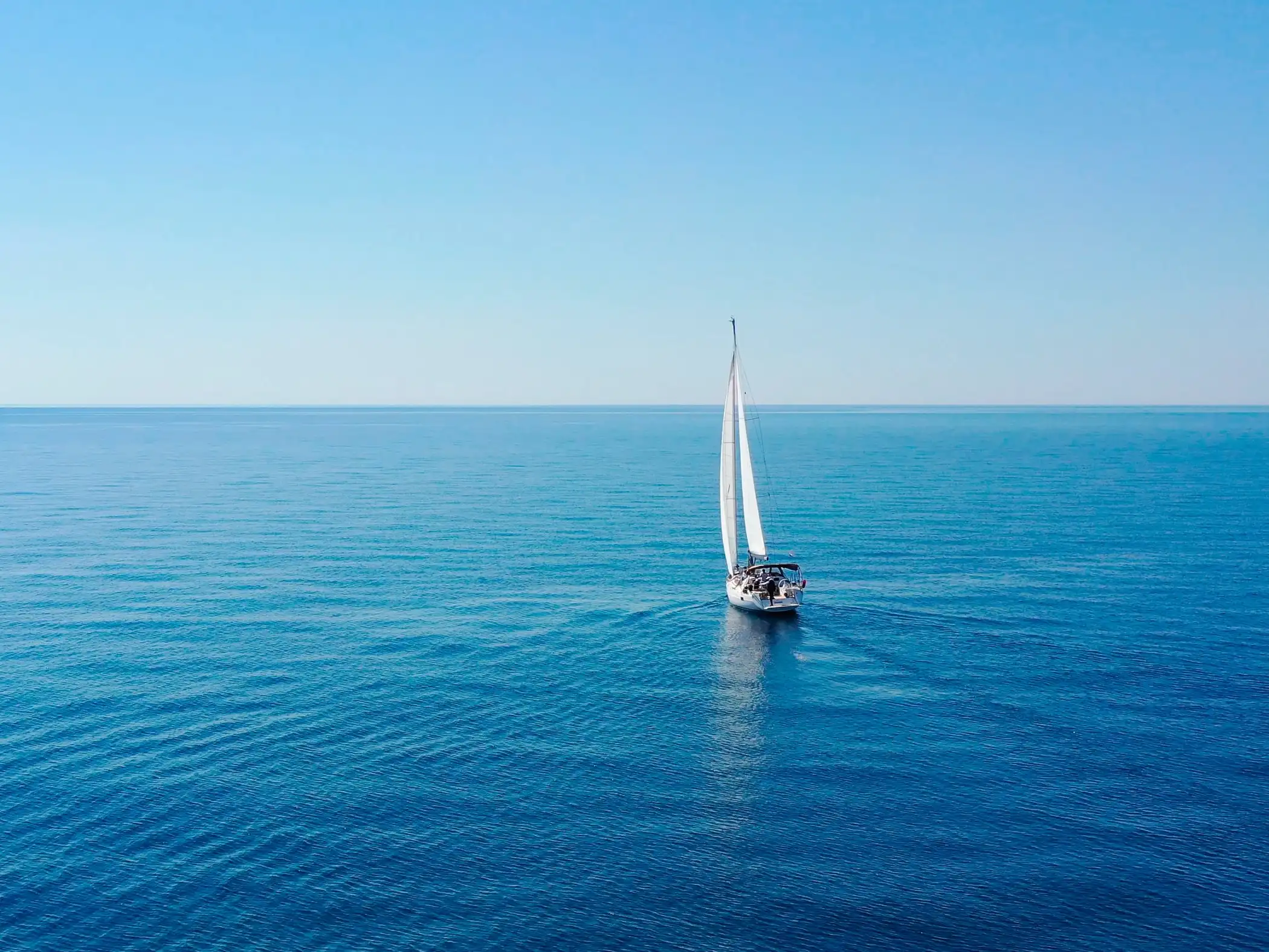 Un voilier sur une mer calme d’un bleu profond, vu du ciel, horizon dégagé